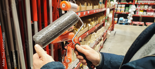 A customer holds a paint tray and brushes while shopping in a hardware store aisle. Concepts of home improvement, DIY renovation, consumer choice, and retail shopping behavior.
