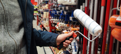 A customer holds a paint tray and brushes while shopping in a hardware store aisle. Concepts of home improvement, DIY renovation, consumer choice, and retail shopping behavior.