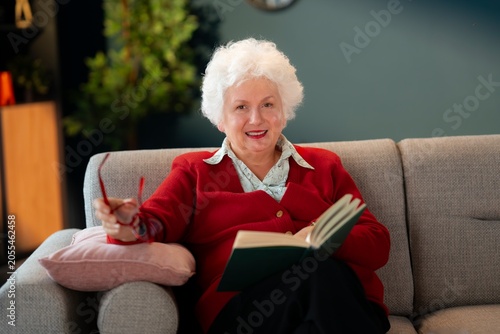 Photos Smiling elderly woman relaxing with book on couch and looking at camera