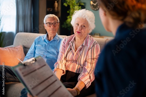 An older couple sits on a couch in a cozy living room. They are having a discussion with a person holding a clipboard. The room is bright and has plants and a clock on the wall.