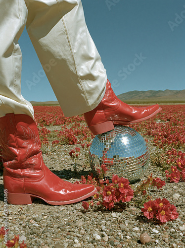 Red cowboy boots rest on a mirrored disco ball in a red wildflower field under a clear blue sky. Fashion editorial concept for footwear branding, lifestyle campaigns and social media.