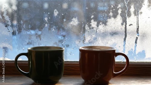 Two steaming mugs of hot beverage on a windowsill with frosty ice patterns on the glass