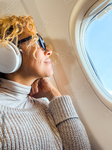 Woman with headphones and sunglasses looking out airplane window, relaxing during a flight in a cozy sweater and turtleneck, enjoying travel, calm, and in-flight comfort