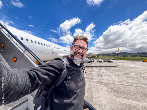 Happy man taking a selfie on airport tarmac while boarding a plane, smiling with backpack under blue sky and clouds, travel, adventure and business trip concept