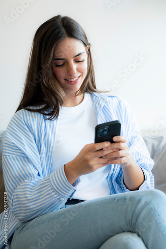 Young woman sitting on a couch smiling while using a smartphone, casually dressed in a striped shirt and jeans, relaxed at home and engaged in texting or browsing social media.