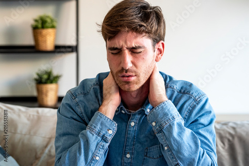 Tableau sur toile Man in denim shirt holding his neck in pain on a sofa at home, eyes closed and w