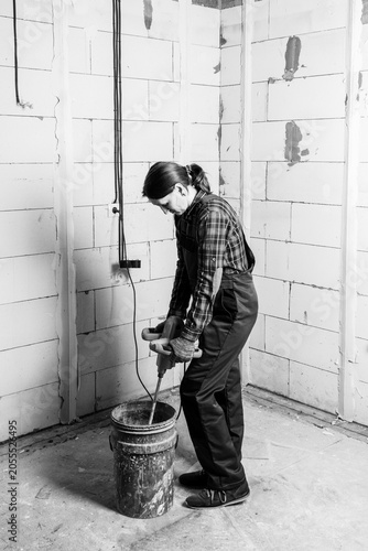 A female plasterer in overalls mixes gypsum mortar for plastering walls in a bucket with an electric mixer black and white photo