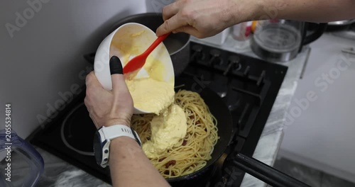 Chef pours sauce over tender spaghetti and golden bacon pieces in frying pan. Experienced cook prepares delicious carbonara dish. Slow motion