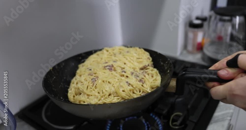 Cook mixes flavorful spaghetti with crispy bacon and creamy sauce by shaking frying pan. Chef prepares carbonara dish in kitchen. Slow motion
