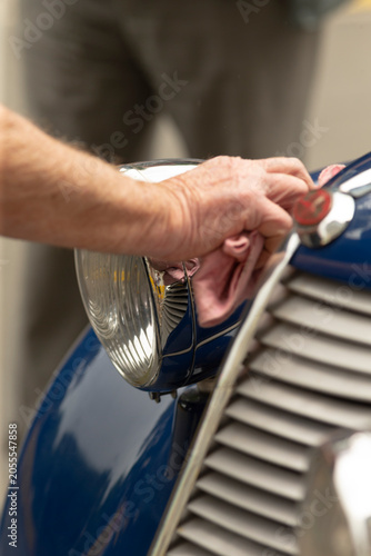 Close view of a person’s hands polishing the chrome headlight and adjacent grille of a glossy blue vintage car with a soft cloth. The scene shows metal