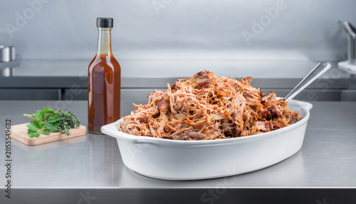 Heaping Pulled Pork in White Ceramic Baking Dish with BBQ Sauce Bottle and Fresh Herbs on Stainless Steel Kitchen Counter