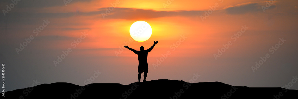 Fototapeta premium A silhouette of a person with arms raised to the sky, embracing the sunset. The person stands atop a hill, with the sun setting behind them, casting a warm, orange glow across the clouds.