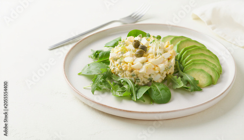 Egg salad tartare with capers and microgreens on mixed greens bed with sliced avocado on white ceramic plate
