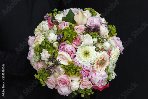 Round luxury bouquet with gerbera daisies, peonies, roses, calla lily and green bell flowers in pink and white, held against black background - bridal floral