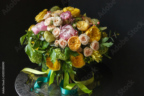 Summer bouquet with yellow garden roses, pink peonies, peach roses and green viburnum in glass vase with yellow and teal ribbons, black background