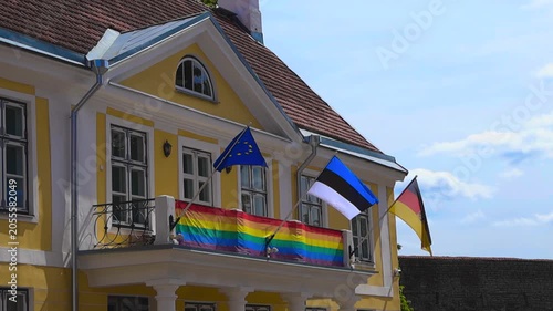 European Union, Estonian, German, and rainbow gay pride flags waving in the wind on the facade of a yellow building, likely an embassy or consulate, showing support for diversity and inclusion