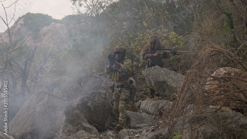 Tableau sur toile A special forces team conducts a tactical operation in a rocky, smoky environment, with a sniper providing covert surveillance from an elevated position