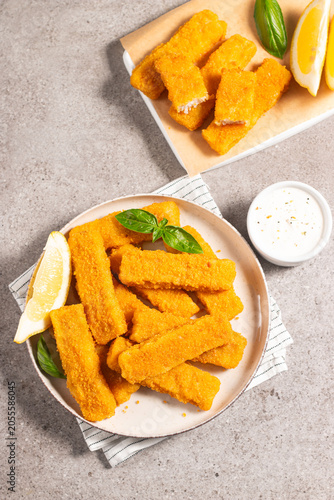 Tasty golden-brown fish fingers served with dip and lemon slices. Crispy breaded fish sticks on light background. 