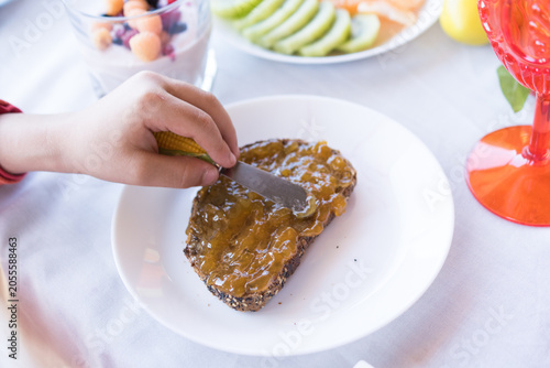 Billede på lærred A close-up shot of a kid’s hand carefully spreading sweet orange marmalade over a slice of multigrain artisan toast on a white plate during breakfast