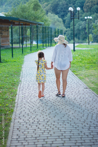 A mother and daughter stroll hand-in-hand down a stone path, enjoying a summer's day outdoors.