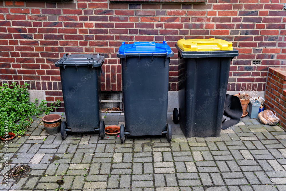 Obraz premium Row of three different sized garbage and recycling plastic containers standing outside on a cobblestone pavement next to a red brick wall.