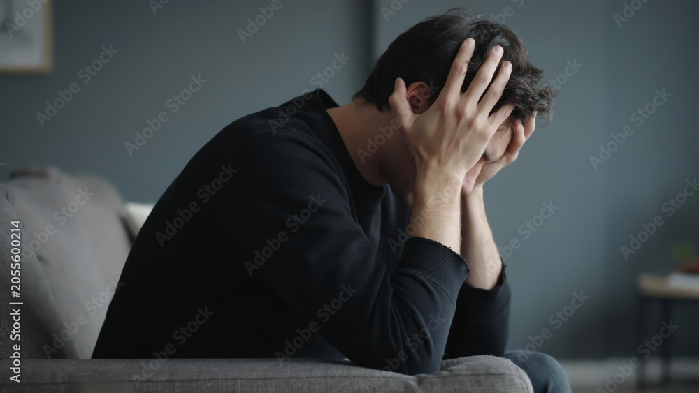 Obraz premium Man holding head in hands sitting on couch in dimly lit room expressing stress and anxiety