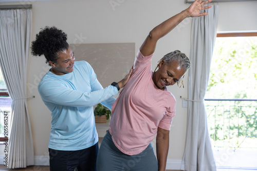 Obraz na plátně Trainer in blue top assisting African American senior woman stretching at home,