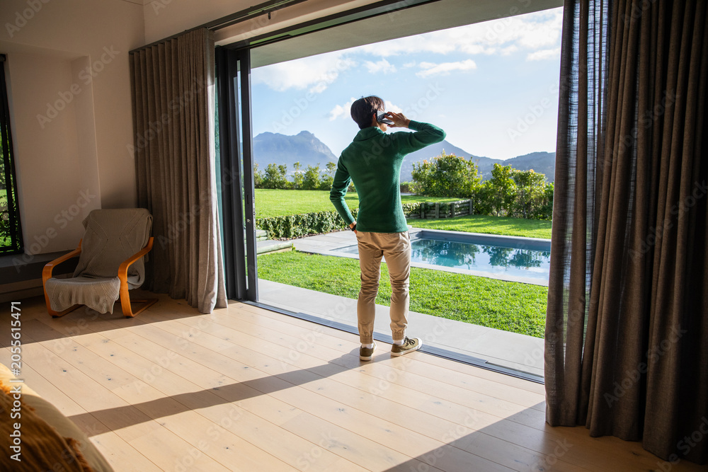 Obraz premium Man is standing by sliding glass door on wood floor, talking on smartphone, pool visible