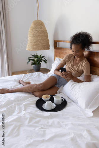 Woman reclining on bed using smartphone with breakfast tray and plant in bright airy bedroom