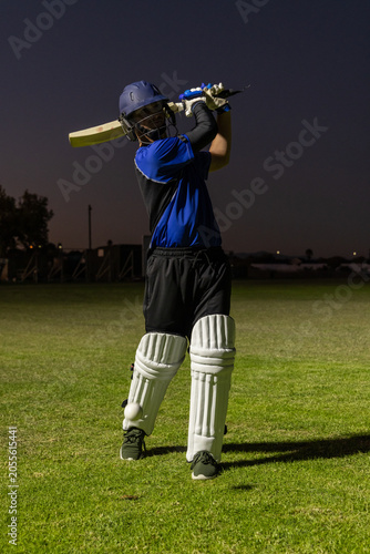 Cricket batsman is holding bat in follow-through on grassy pitch at dusk with ball near pad