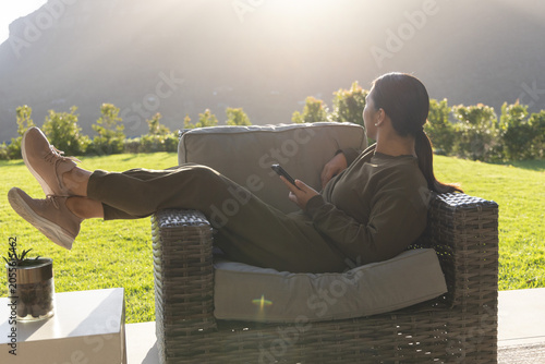 Woman reclining on woven armchair on patio holding phone and watching sunlit lawn with lens flare
