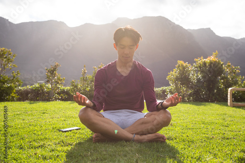 Asian adult man meditating cross-legged on grassy lawn with smartphone left, anklet showing