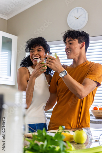 Diverse couple clinking clear glasses and laughing at counter after blender finishing green juice