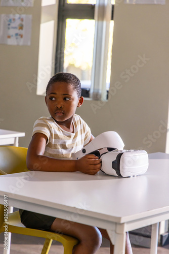 African American schoolboy sitting on yellow chair at desk holding white VR headset in classroom