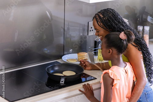 African American mom and daughter cooking pancakes at stove using spatula and pan, copy space