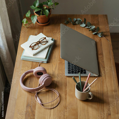 Laptop sits open on a wooden desk surrounded by a notebook, headphones, and a small potted plant.