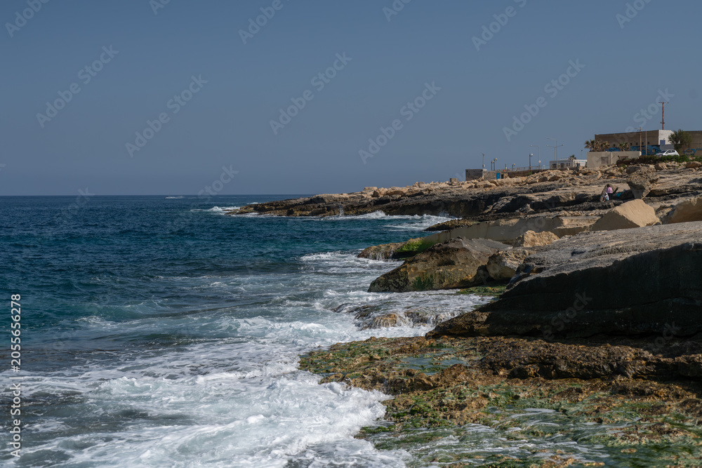 Fototapeta premium Rugged limestone coastline in Malta with turquoise Mediterranean waves breaking over algae covered rocks, capturing dramatic surf, coastal erosion, and natural seascape under a clear blue sky.