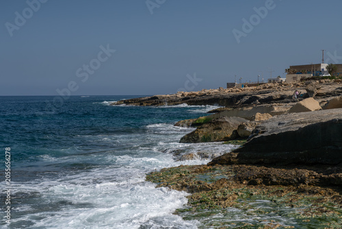 Rugged limestone coastline in Malta with turquoise Mediterranean waves breaking over algae covered rocks, capturing dramatic surf, coastal erosion, and natural seascape under a clear blue sky.