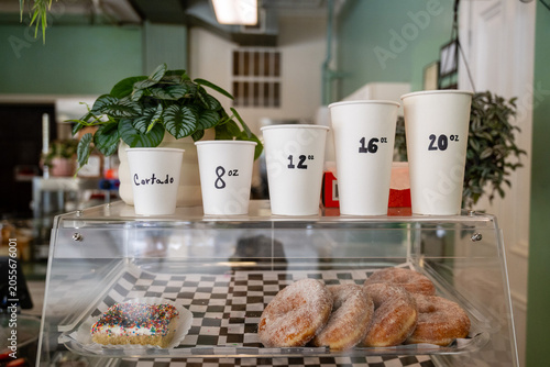 Close up of a coffee shop counter with stacked mugs showing drink sizes and prices next to green potted plants.