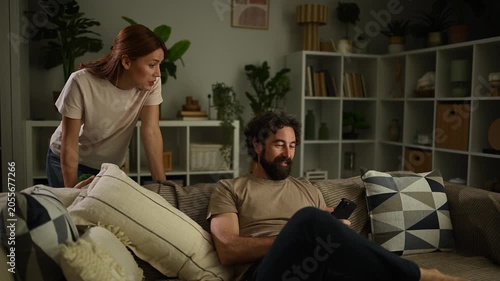 Angry wife scolding lazy husband using his phone on the sofa, causing a relationship conflict and argument in the living room