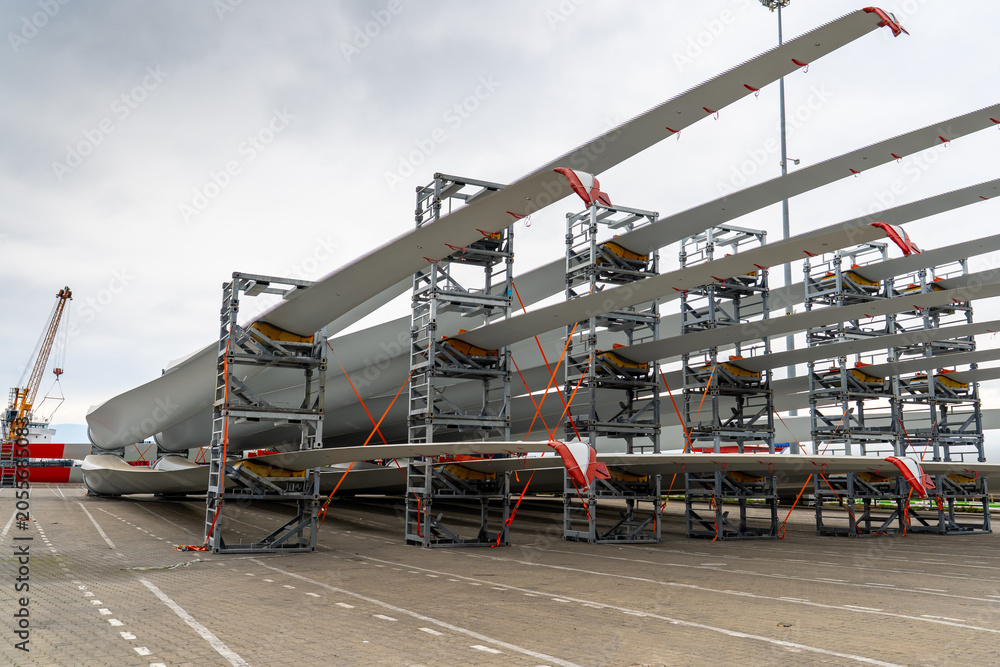 Naklejka premium Wind turbine blades stored in transport racks at a port logistics terminal. Renewable energy components prepared for shipment, heavy cargo handling and wind farm infrastructure.