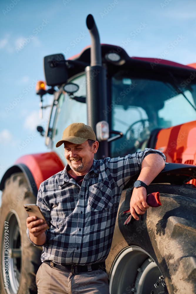 Naklejka premium Happy agriculturist using cell phone while leaning on tractor.