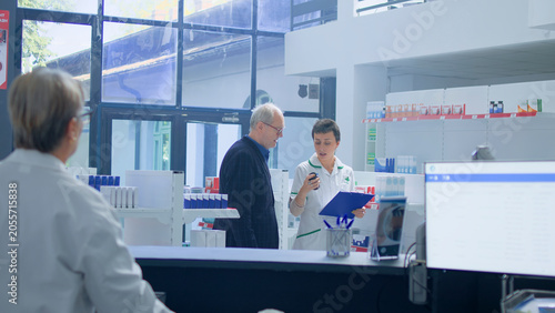 Bild auf Leinwand Elderly man in need of prescription medicaments informations in apothecary, requesting assistance from cashier who redirects him to qualified chemist checking pharmaceutical products inventory
