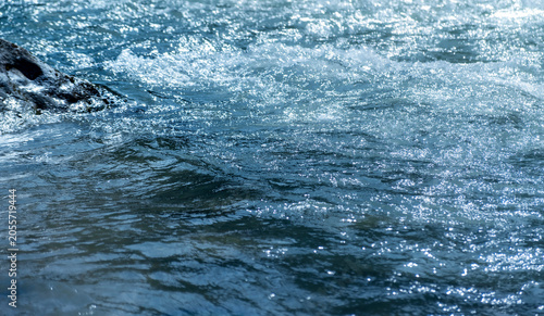 Close-up of rushing river surface with splashing ripples. River rushing over rocks. The cool water creates white splashes and crystalline droplets in bright sunlight. Evoking purity or natural energy.