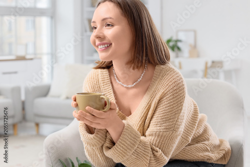 Happy young woman with cup of coffee sitting in armchair at home