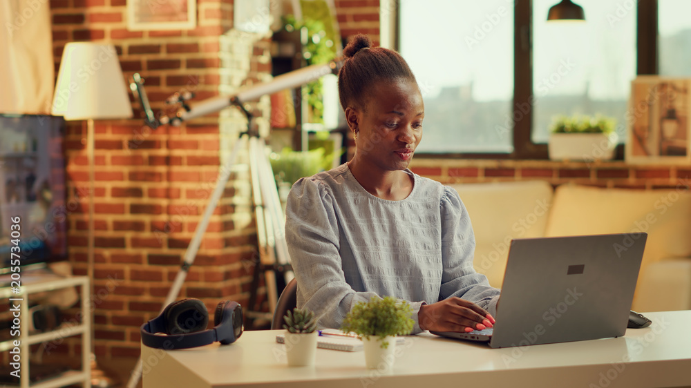 Fototapeta premium African American girl operates from home in the glow of sunset, publishing personal website article. Teleworker seeking to build online freelance business while refining abilities. Tripod shot.