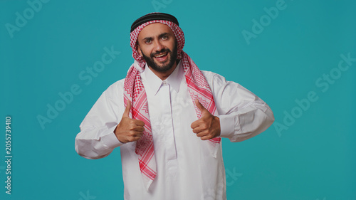 Person wearing customary thobe and kufiyah seems joyful, exhibiting genuine muslim beliefs and practices. Middle eastern guy with distinctive culture, dress and structured scarf.