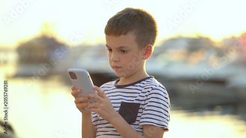 Foto An eight-year-old boy sits on the shore and plays the phone against the backdrop