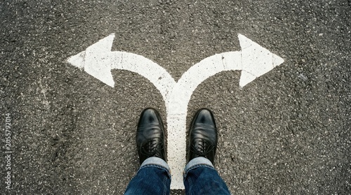 Business person standing on asphalt road wearing black leather shoes looking at white painted arrows pointing in two opposite directions