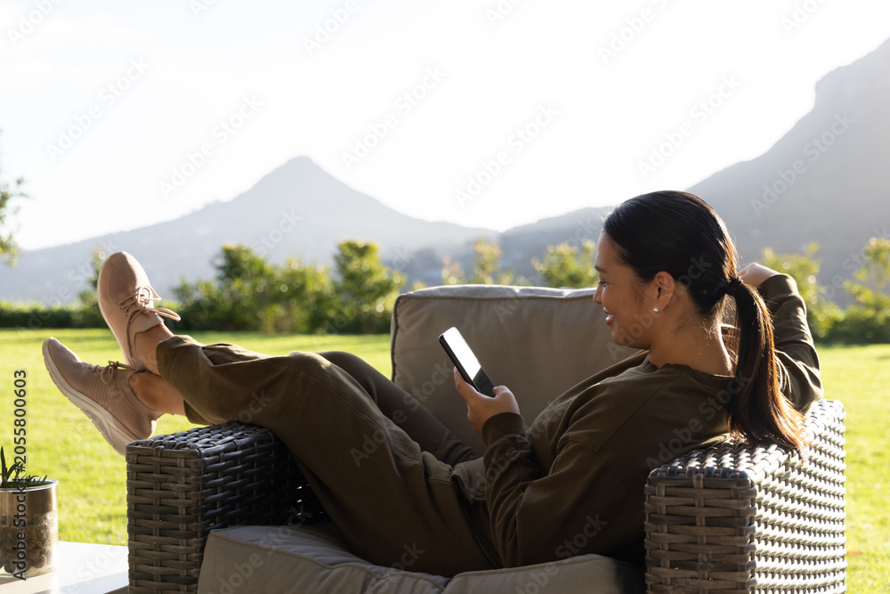 Fototapeta premium Wicker armchair holding olive loungewear and smartphone on patio, lawn and conical mountain beyond